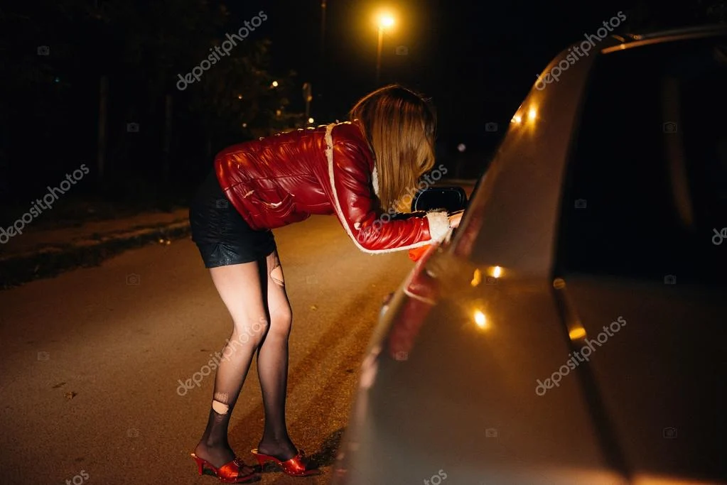 Woman in a red jacket leans over a car at night on a dimly lit street.