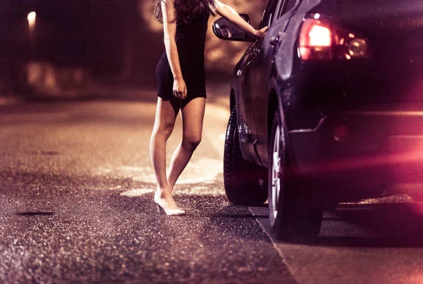 woman in a black dress standing by a car on a dimly lit street at night