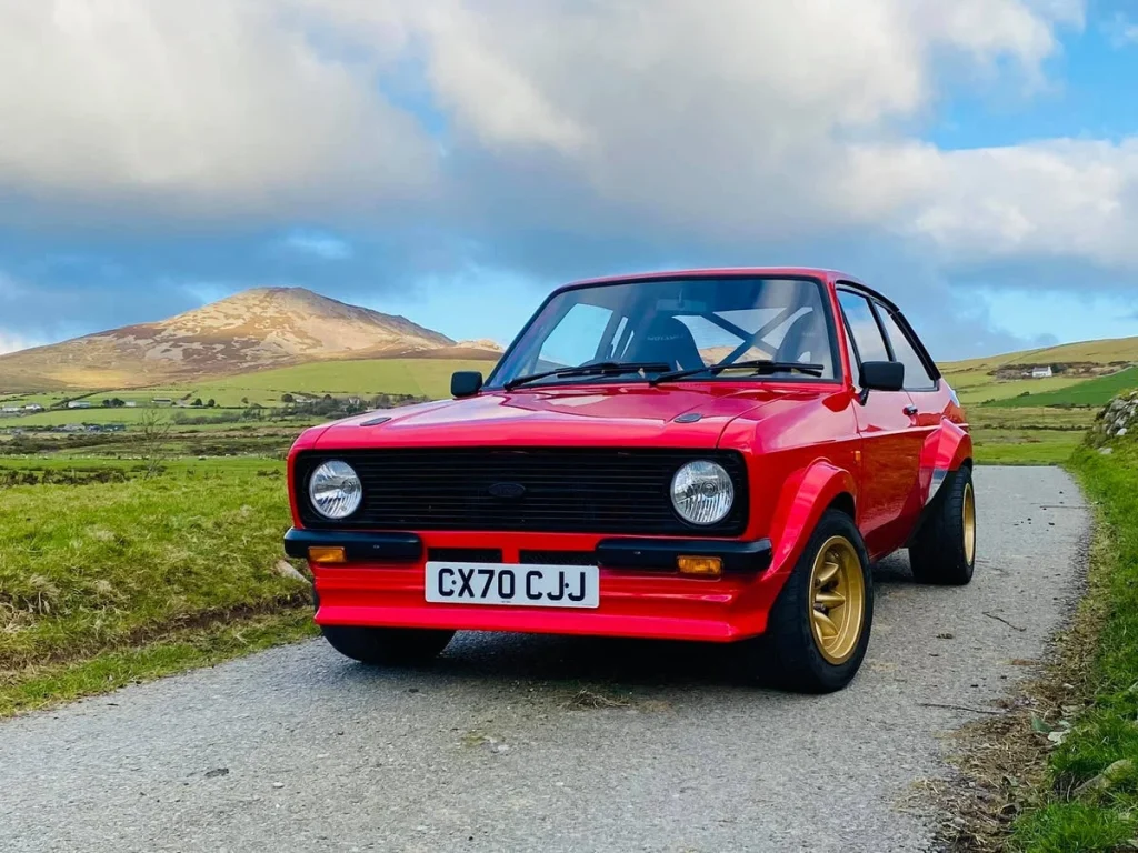 Red 1980 Ford Escort rally car parked on a scenic road with hills in the background.