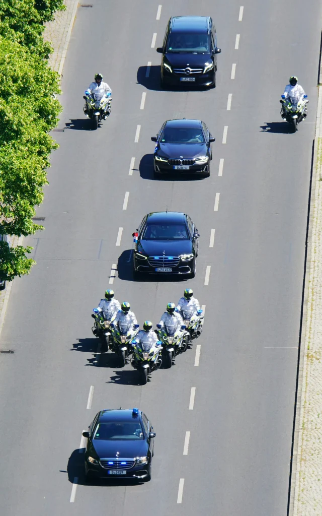 Motorcade with police motorcycles and black vehicles on a city street, flanked by trees.