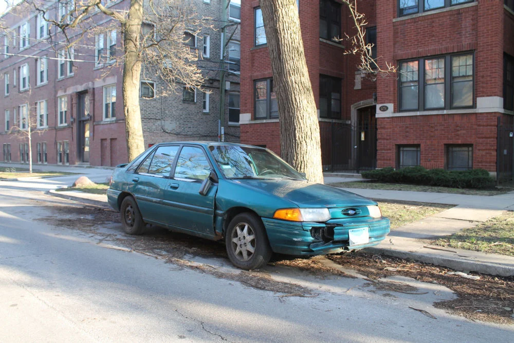 A damaged green 1995 Ford Escort parked on a city street beside a tree and brick buildings.