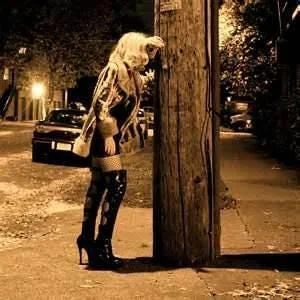 A woman in high heels leans against a street pole at night, with dim streetlights illuminating the scene.