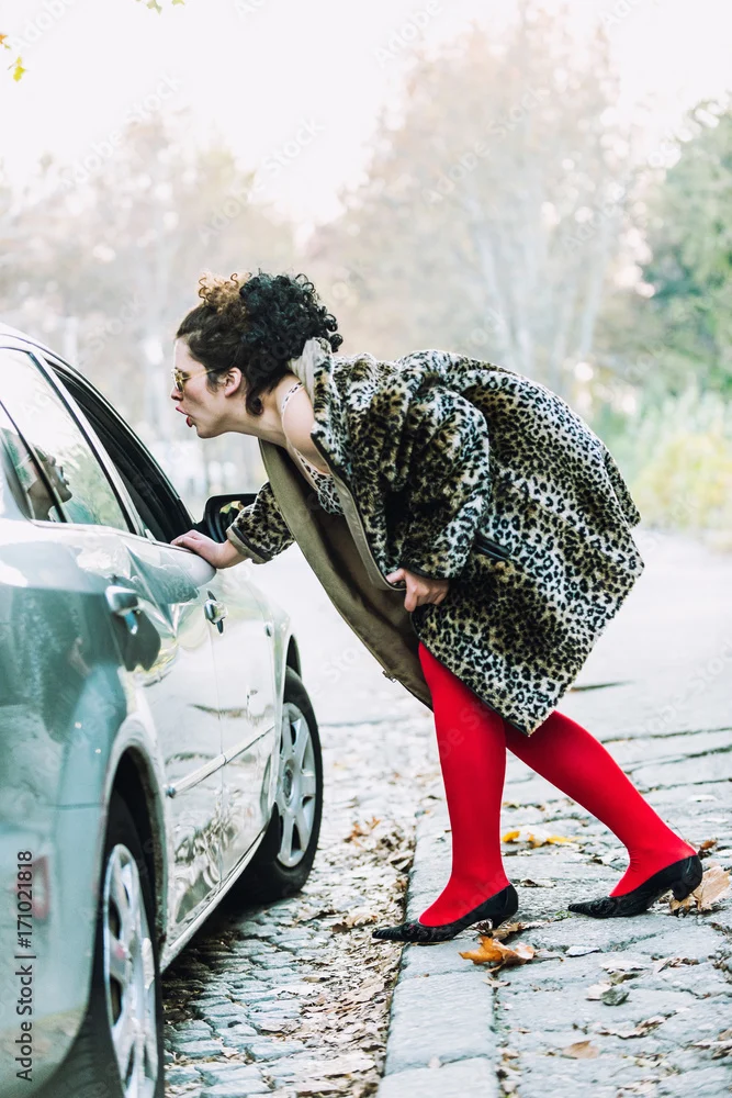 A woman in a leopard print coat and red tights leans toward a car window on a cobblestone street.