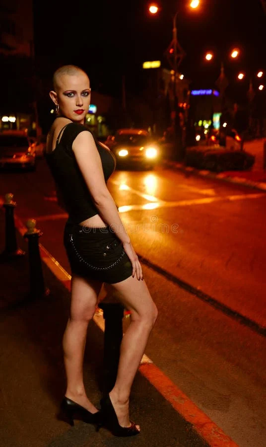 woman with a shaved head in a black outfit poses on a city street at night, illuminated by streetlights and car headlights
