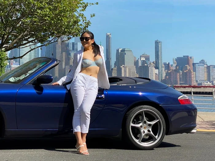 Woman in a bikini top and white pants poses by a blue convertible with a city skyline in the background.